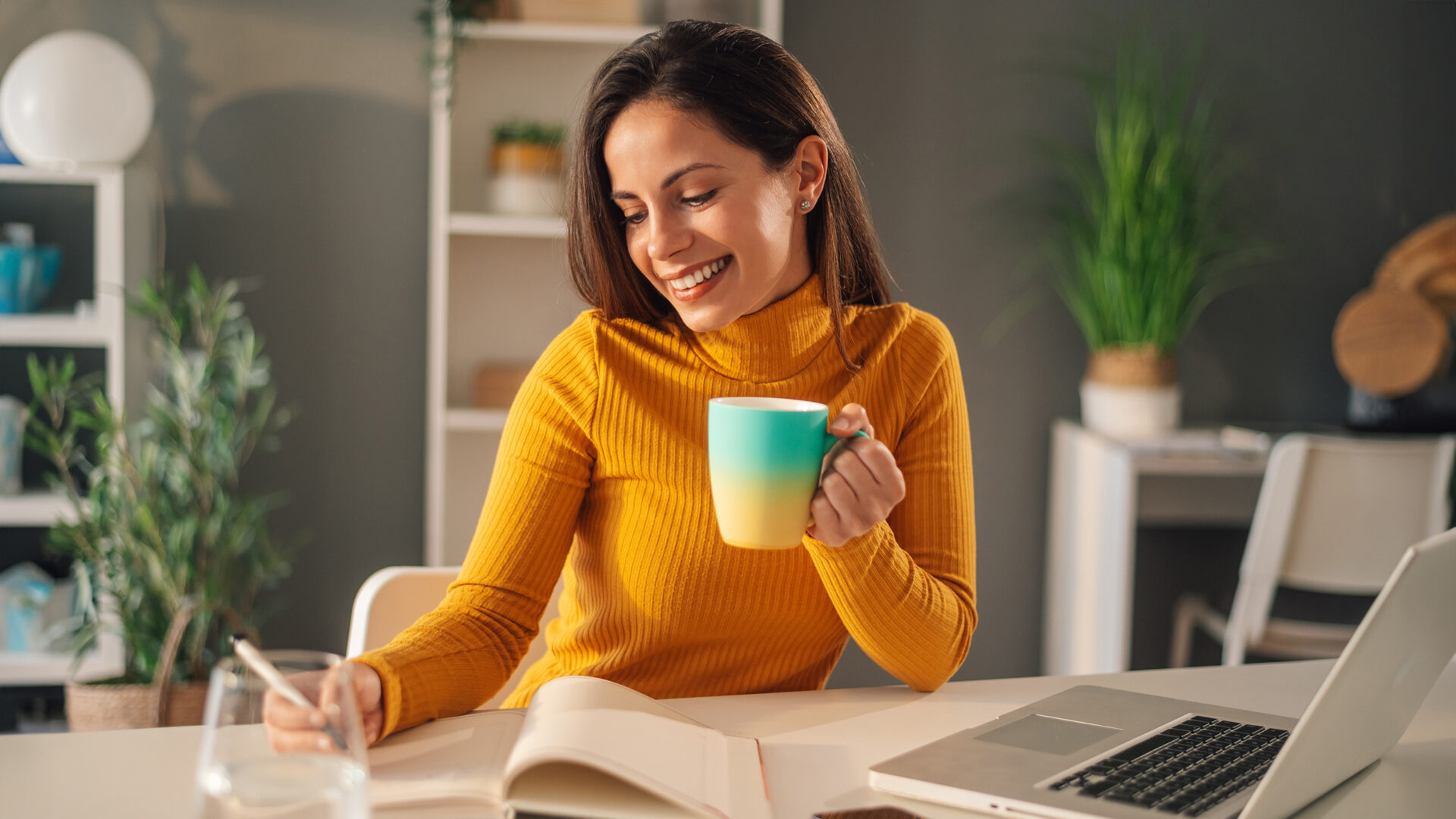 Vrouw met kop koffie aan tafel, voor haar staat een laptop en notitieblok