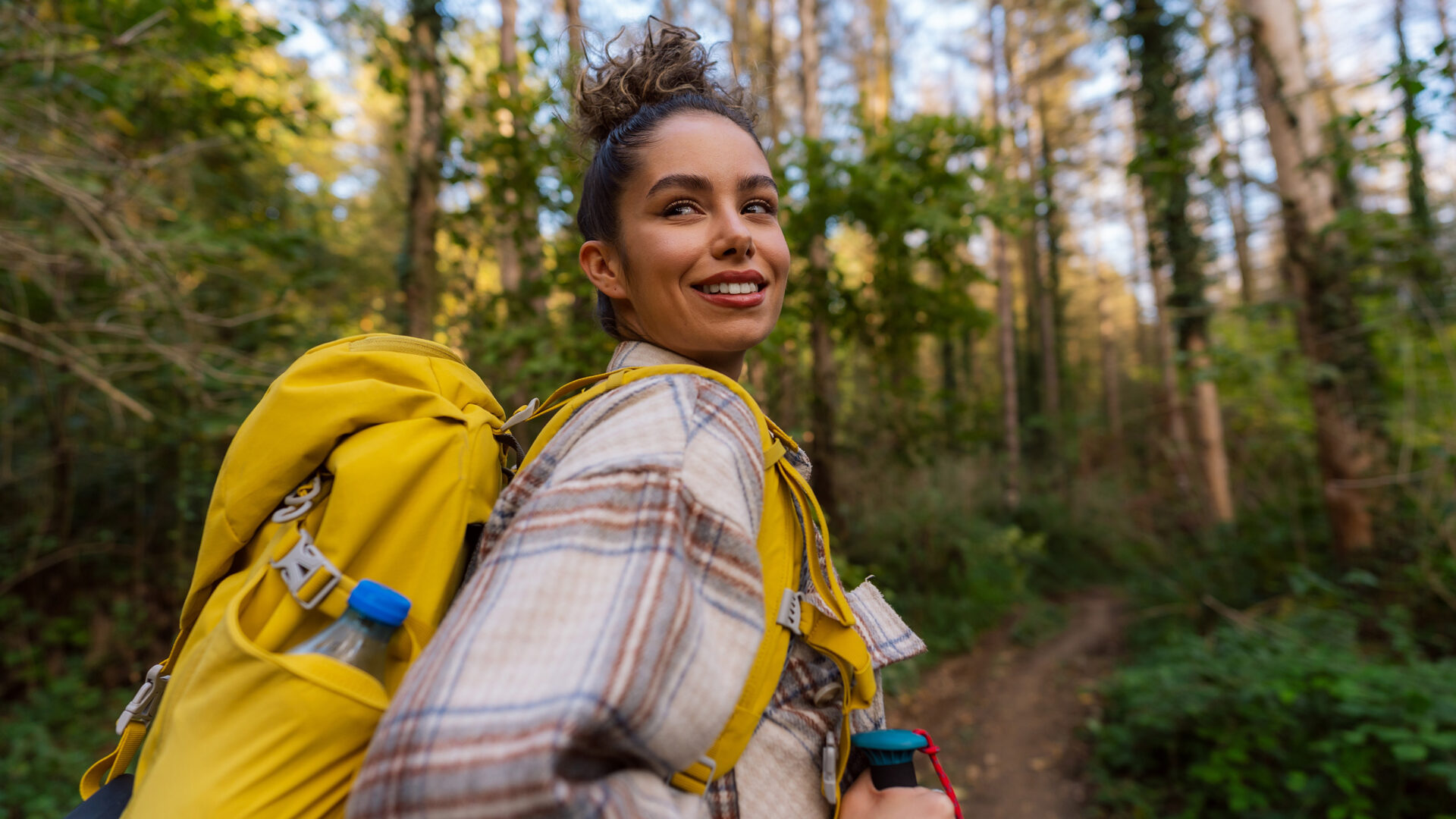 Vrouw met gele rugzak in het bos, kijkt over haar schouder naar de camera