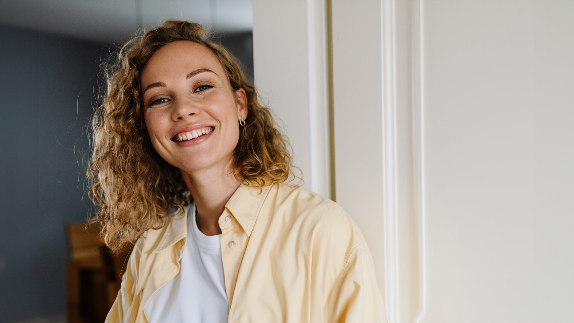 Vrouw met wit shirt en gele blouse leunt lachend tegen deurpost