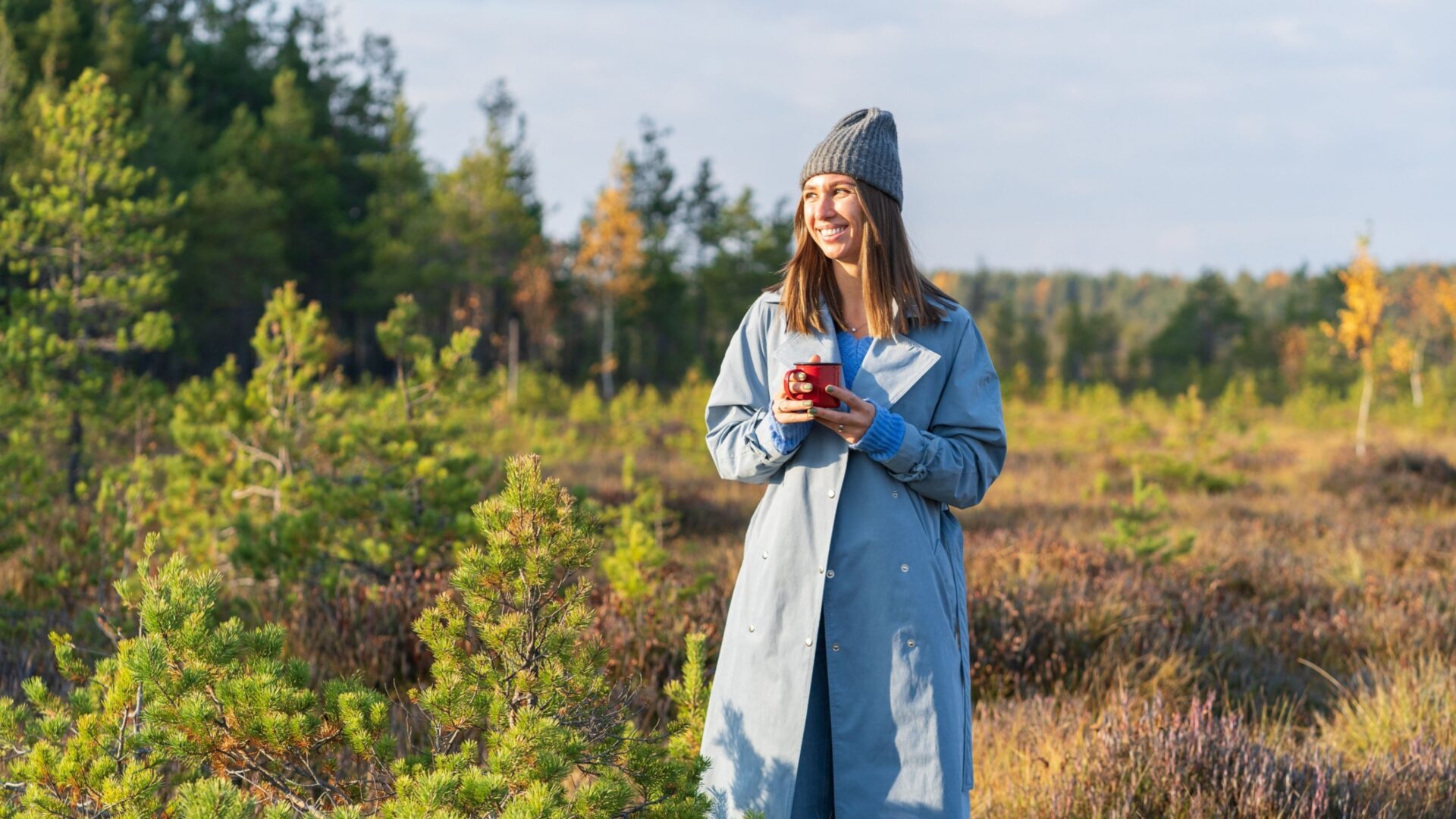 Vrouw met een mok, regenjas en een muts op op een openvlakte in het bos