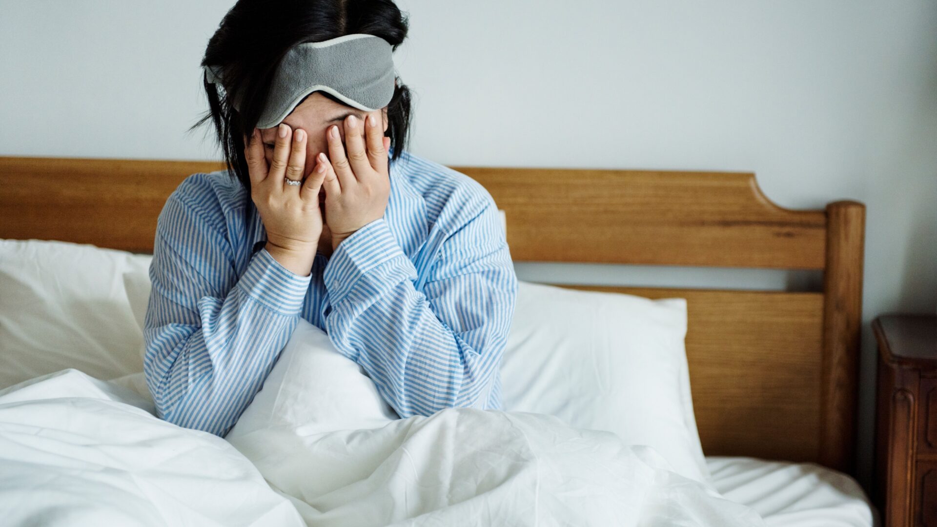 Vrouw rechtop in bed met slaapmasker en handen voor haar gezicht